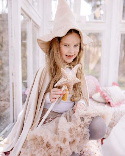 Mała dziewczynka w beżowym kapeluszu czarownicy i pelerynie, trzymająca różdżkę w kształcie różowej, błyszczącej gwiazdy, siedzi na łóżku w jasnym pokoju z dużymi oknami.