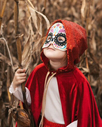 Dziewczynka w czerwonym pelerynie z kapturem i kolorowej masce trzyma suchy kij stojąc pośród uschniętych liści kukurydzy.