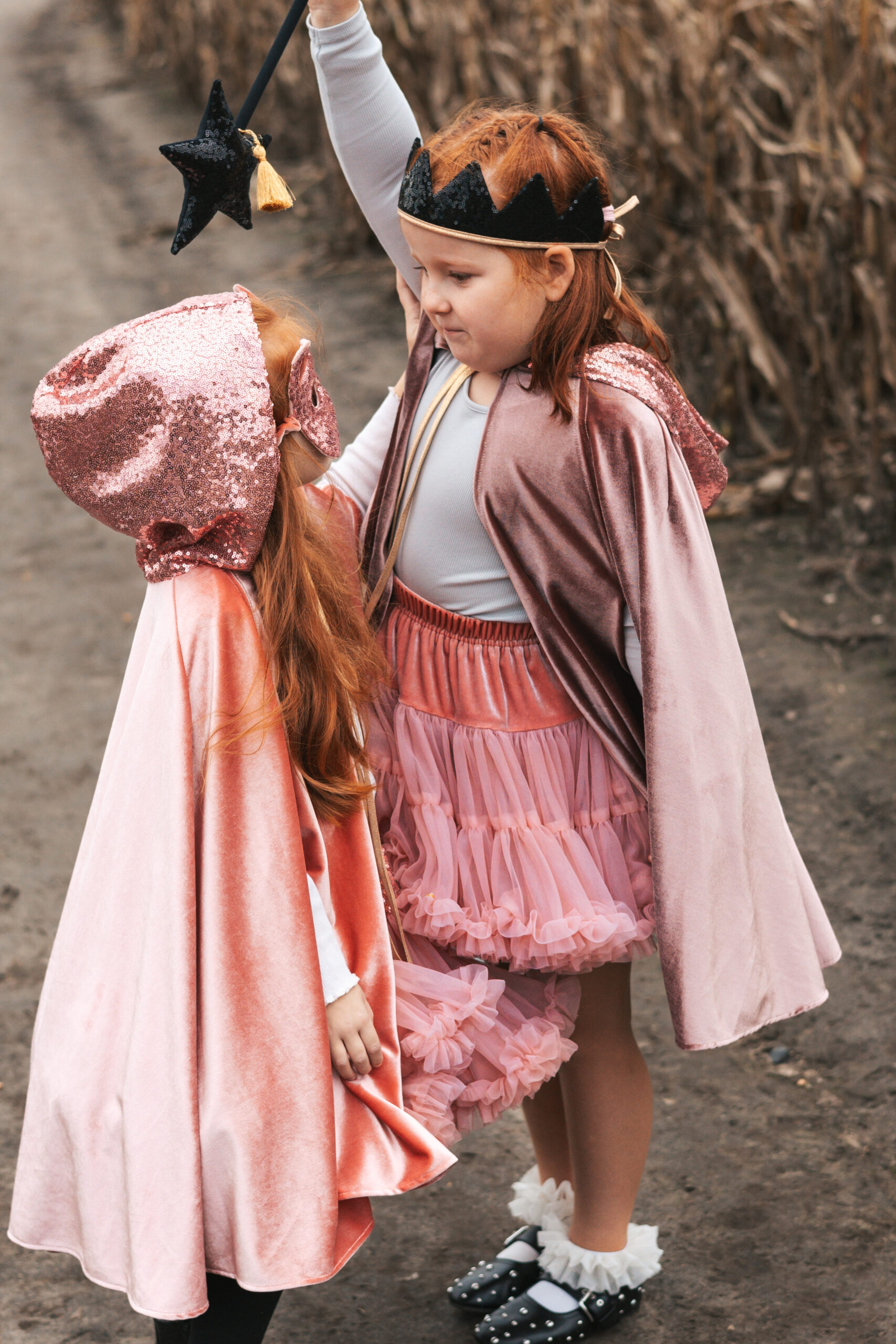 Dwie dziewczynki w różowych, połyskujących pelerynach i kostiumach, jedna z koroną i różową tiulową spódniczką, druga z kapturem i maską, stoją na drodze obok pola kukurydzy, starsza trzyma czarodziejski różdżkowy różdżkę podniesioną nad młodszą.