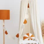Biały baldachim nad dziecięcym łóżeczkiem z pomarańczowo-białą poduszką i lampą w kolorze pomarańczowym obok