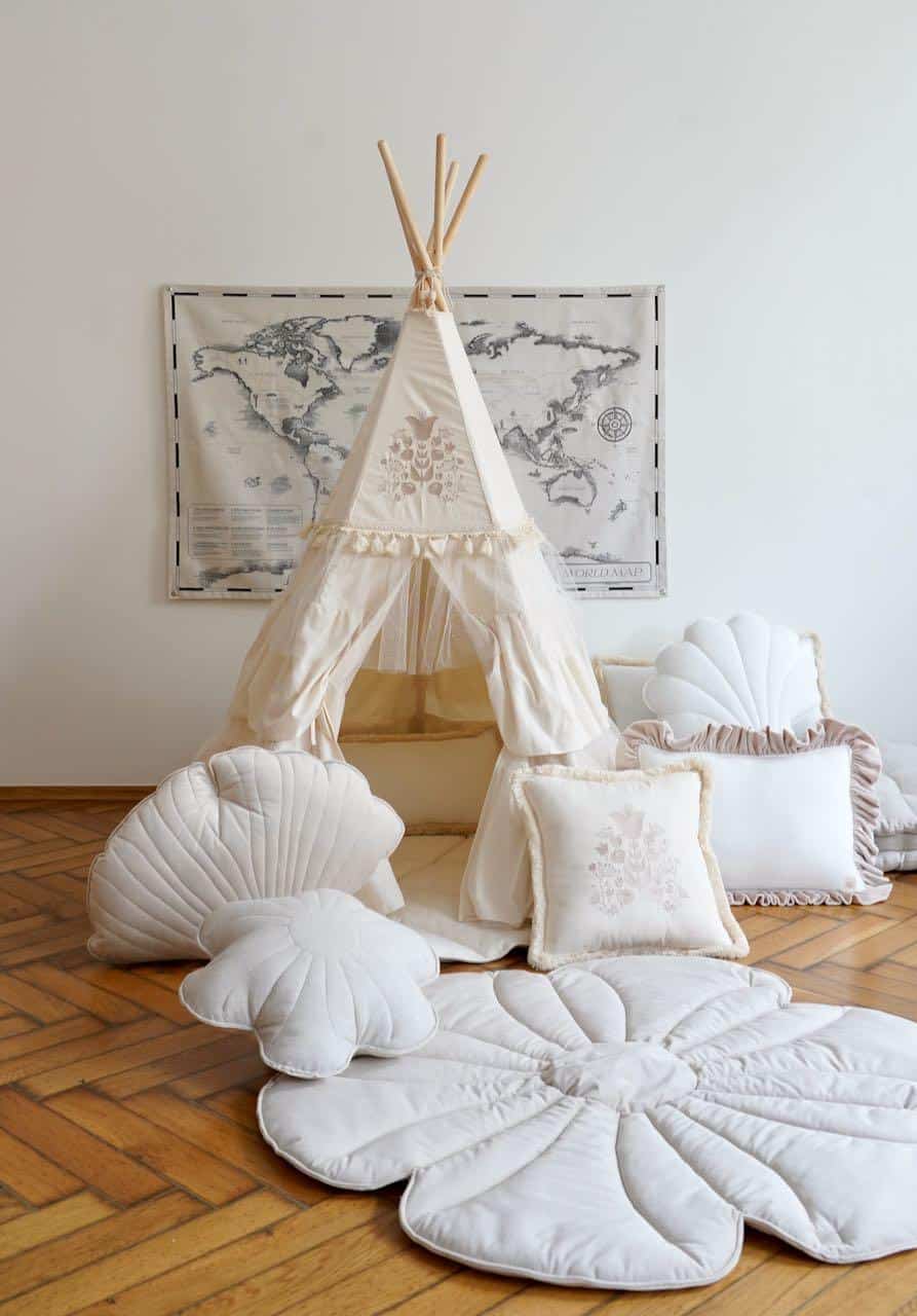 Dziecięcy namiot tipi z beżowej tkaniny, ozdobiony dekoracyjnymi poduszkami i matą w kształcie kwiatów