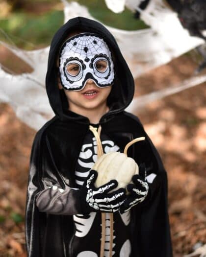 Dziecko w kostiumie szkieletu z maską i czarną peleryną trzymające białą dynię na Halloween