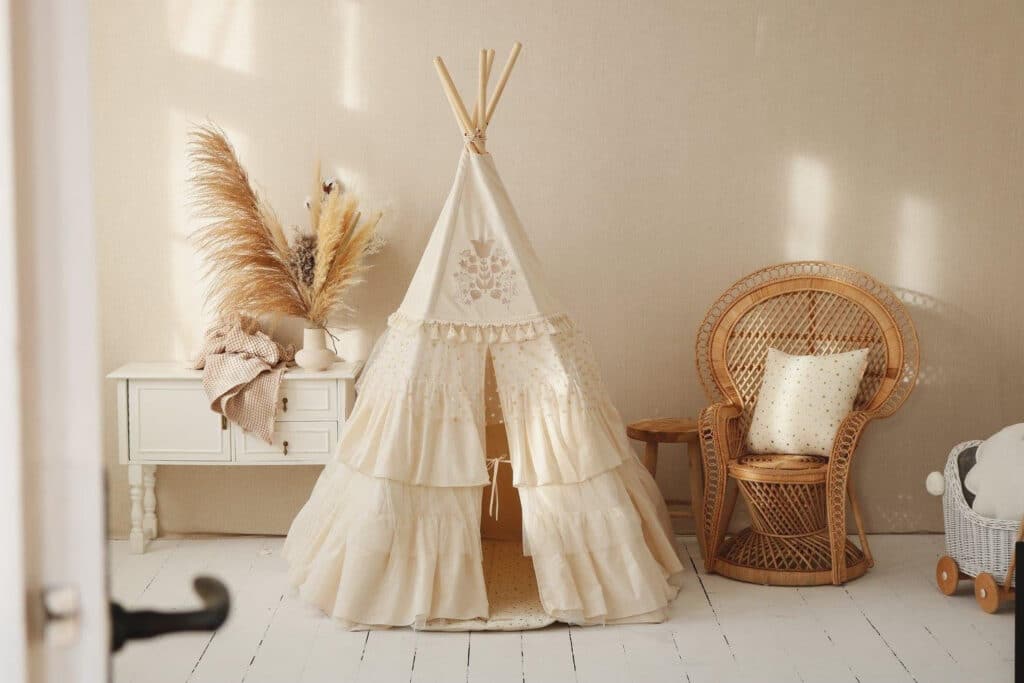 Przytulny kącik dziecięcy z beżowym tipi, wiklinowym fotelem i białą szafką z dekoracjami