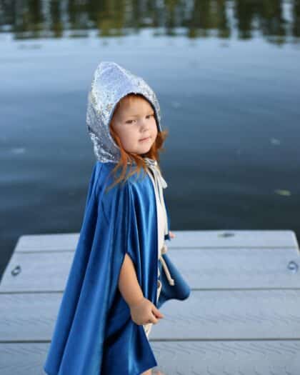 Mała dziewczynka w niebieskim płaszczu z cekinowym kapturem stoi nad wodą na pomoście na tle jeziora