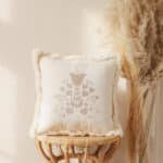 Beżowa poduszka z haftowanym kwiatowym wzorem na wiklinowym stołku obok wysokiej wazonu z trawami pampasowymi