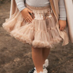 Dziecko w beżowej tiulowej spódnicy z cekinowym pasem, szarej bluzce i beżowym płaszczu, stojące na ziemi, noszące białe skarpetki z falbanami i czarne buty z ozdobami.