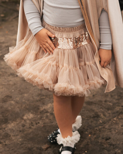 Dziecko w beżowej tiulowej spódnicy z cekinowym pasem, szarej bluzce i beżowym płaszczu, stojące na ziemi, noszące białe skarpetki z falbanami i czarne buty z ozdobami.