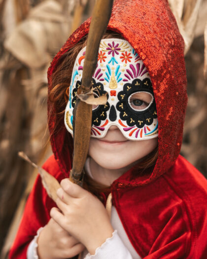 Dziecko w czerwonym, cekinowym kapturze i kolorowej masce z motywem kwiatu, trzymające suchą gałąź na tle suchych liści kukurydzy.