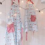 Komplet fartuchów kuchennych z motywem przyborów do gotowania, z czerwonymi kieszeniami w kratkę, zawieszonych na białej ścianie.