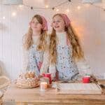 Dwie dziewczynki w fartuszkach i chustach stoją przy stole z piernikowym domkiem i świątecznym wystrojem.