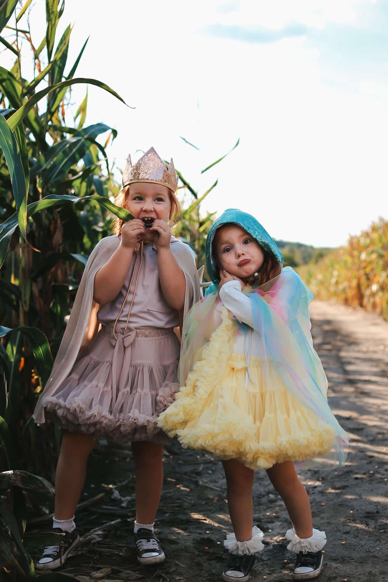 Dwie małe dziewczynki w kolorowych sukienkach i pelerynach stoją na ścieżce między kukurydzą na polu
