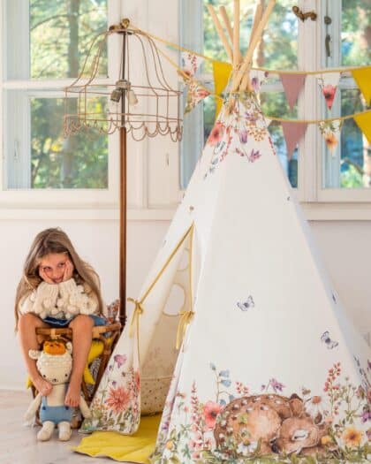 Dziewczynka siedzi przy tipi z motywem jelenia i kwiatów w jasnym, przytulnym pokoju z girlandą.