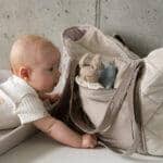 Niemowlę w białym pajacyku z wzorem ciekawie patrzy na pluszowego króliczka w beżowej torbie na stole