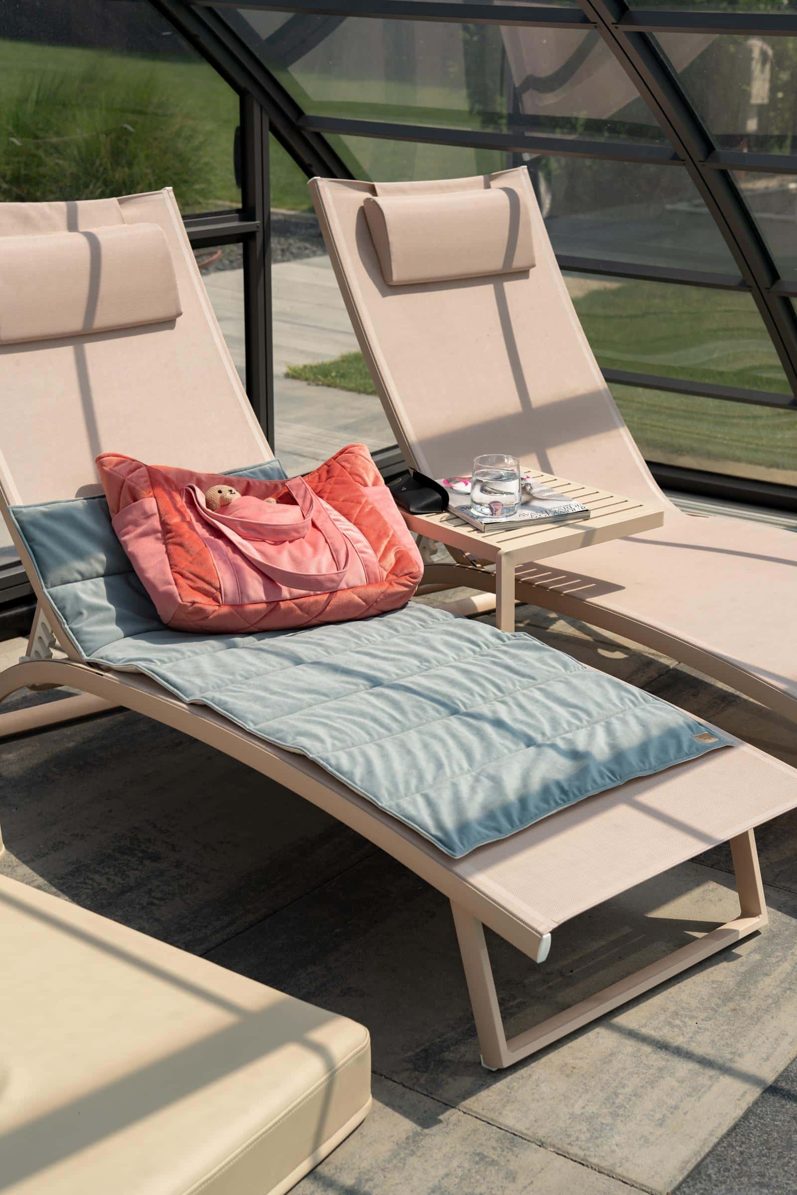 Dwa beżowe leżaki w oranżerii, na jednym niebieska mata i różowa torba z pluszakiem, na drugim stolik z szklanką wody