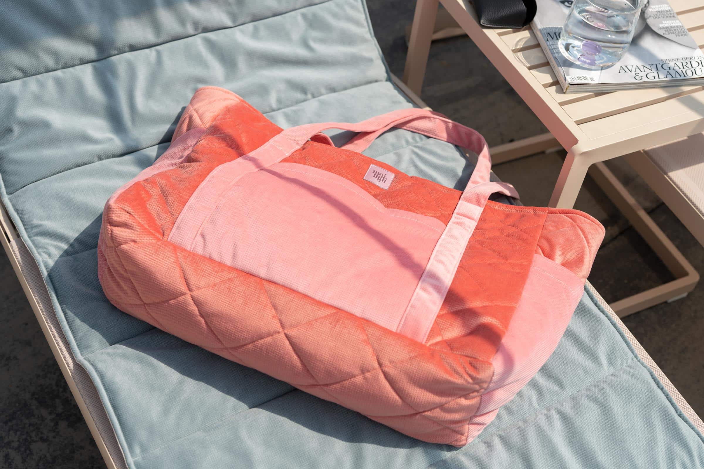 Różowa pikowana torba na miękkim niebieskim leżaku obok stolika z gazetami i szklanką wody