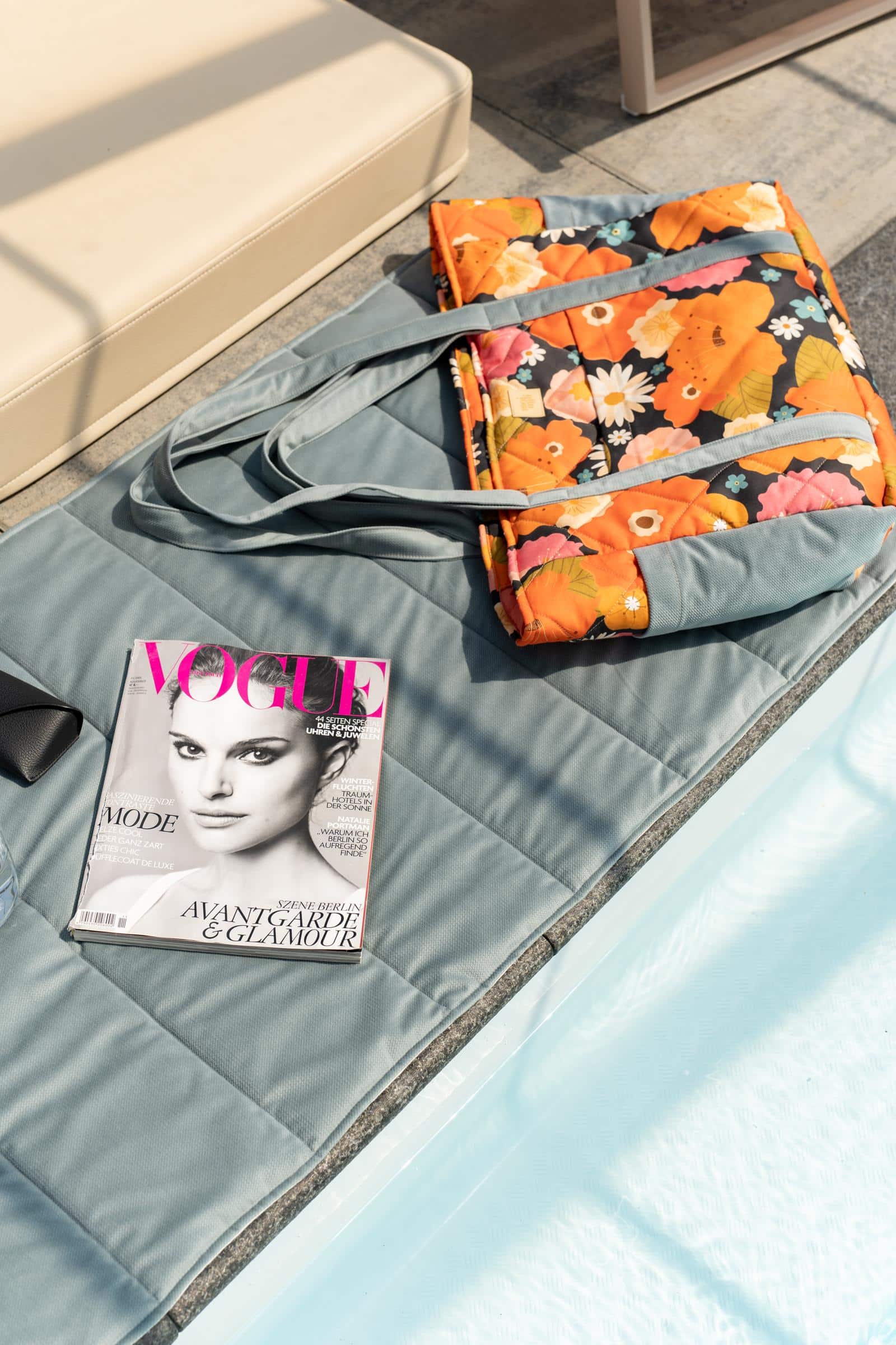 Leżąca przy basenie niebieska mata z kolorową torbą w kwiaty i magazynem Vogue na niej.