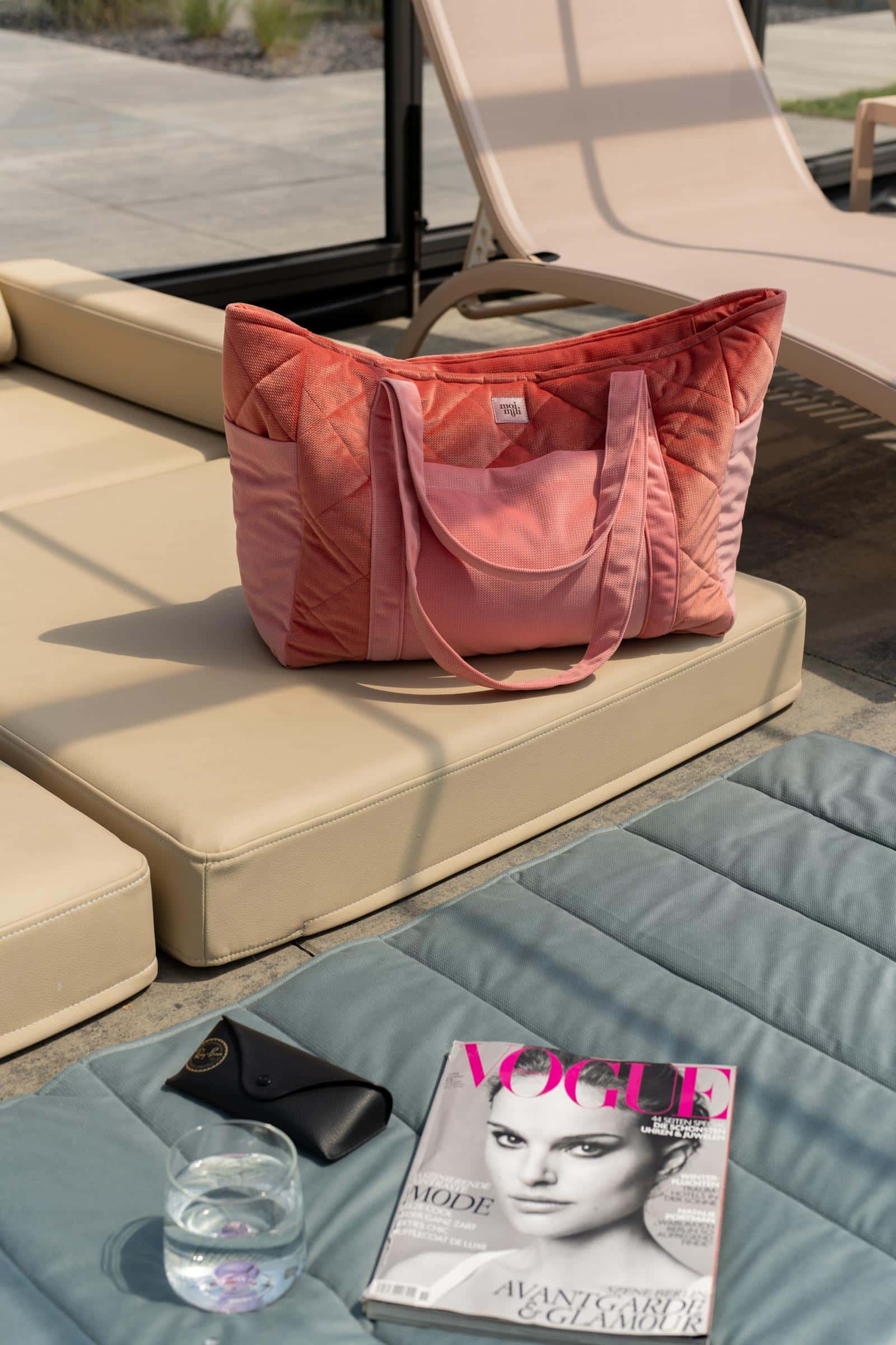 Różowa pikowana torba na beżowej sofie, przy niej leży magazyn Vogue, okulary i szklanka wody na szarej poduszce