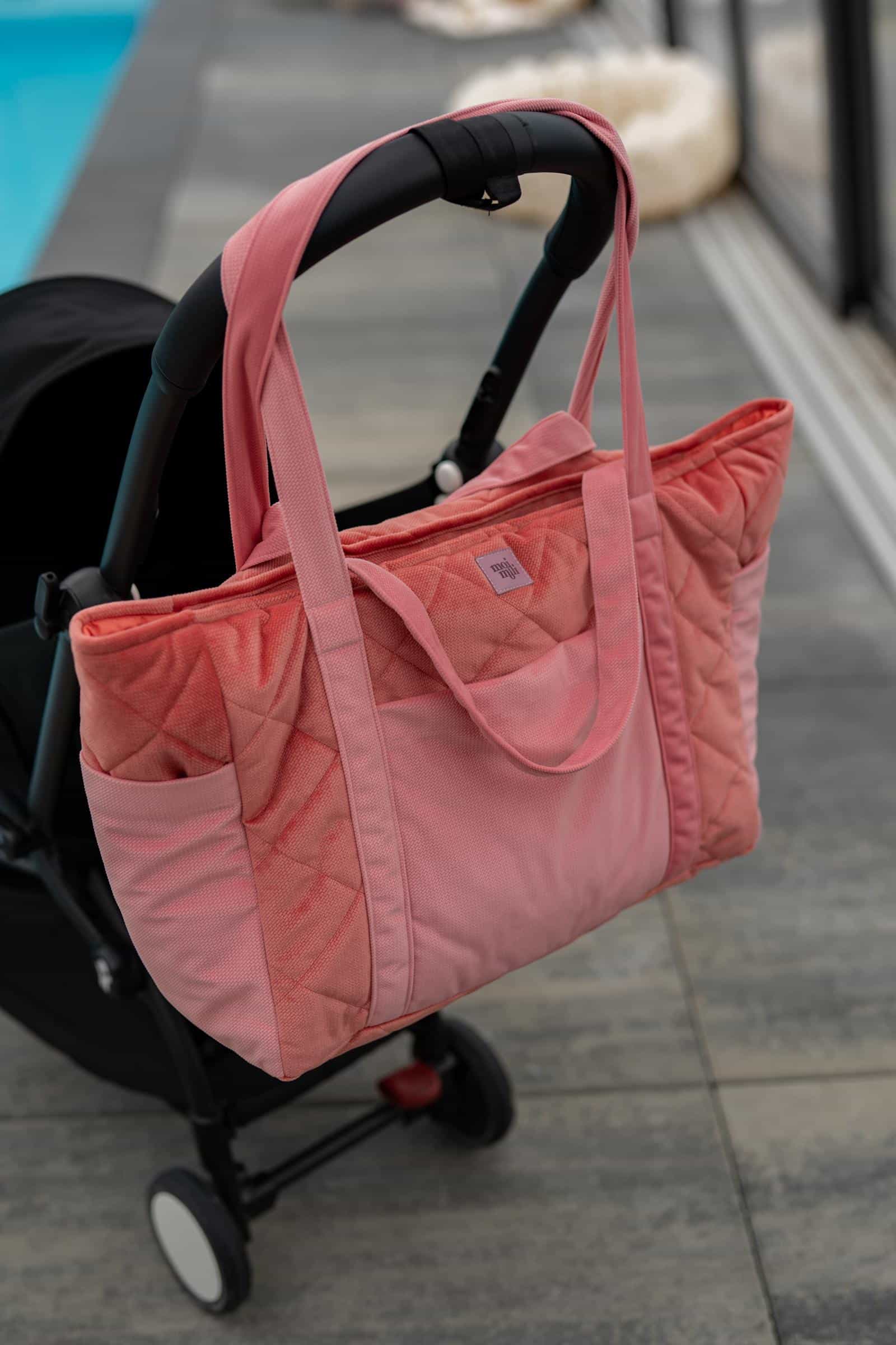 Różowa torba zawieszona na czarnej rączce wózka dziecięcego stojącego na szarej posadzce.