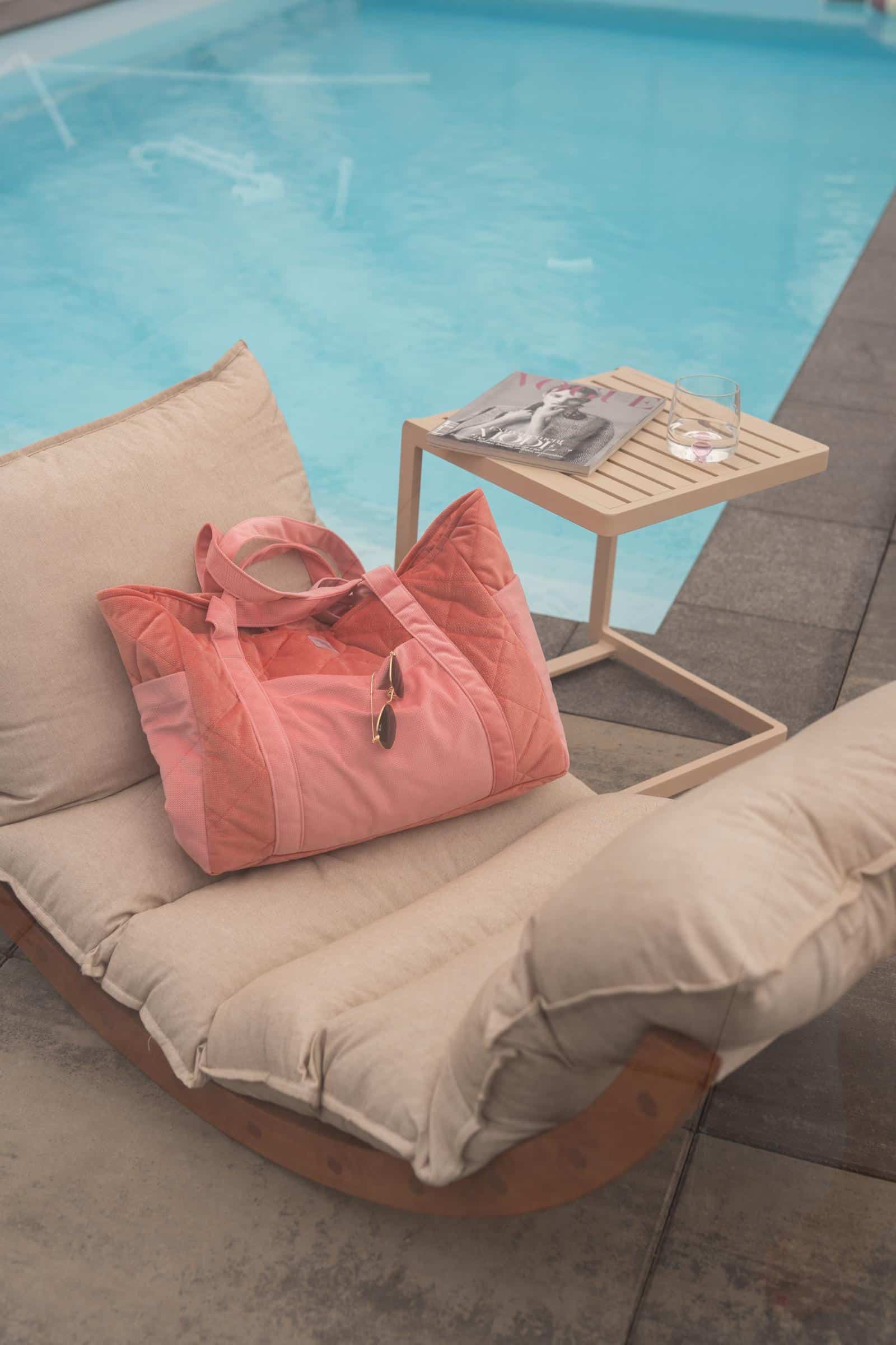 Beżowy fotel ogrodowy z różową torbą i okularami, obok stolik z magazynem i szklanką, przy basenie