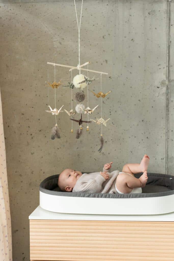 Niemowlę leżące na przewijaku z zawieszonym nad nim dekoracyjnym karuzelą z papierowymi żurawiami i piórami.