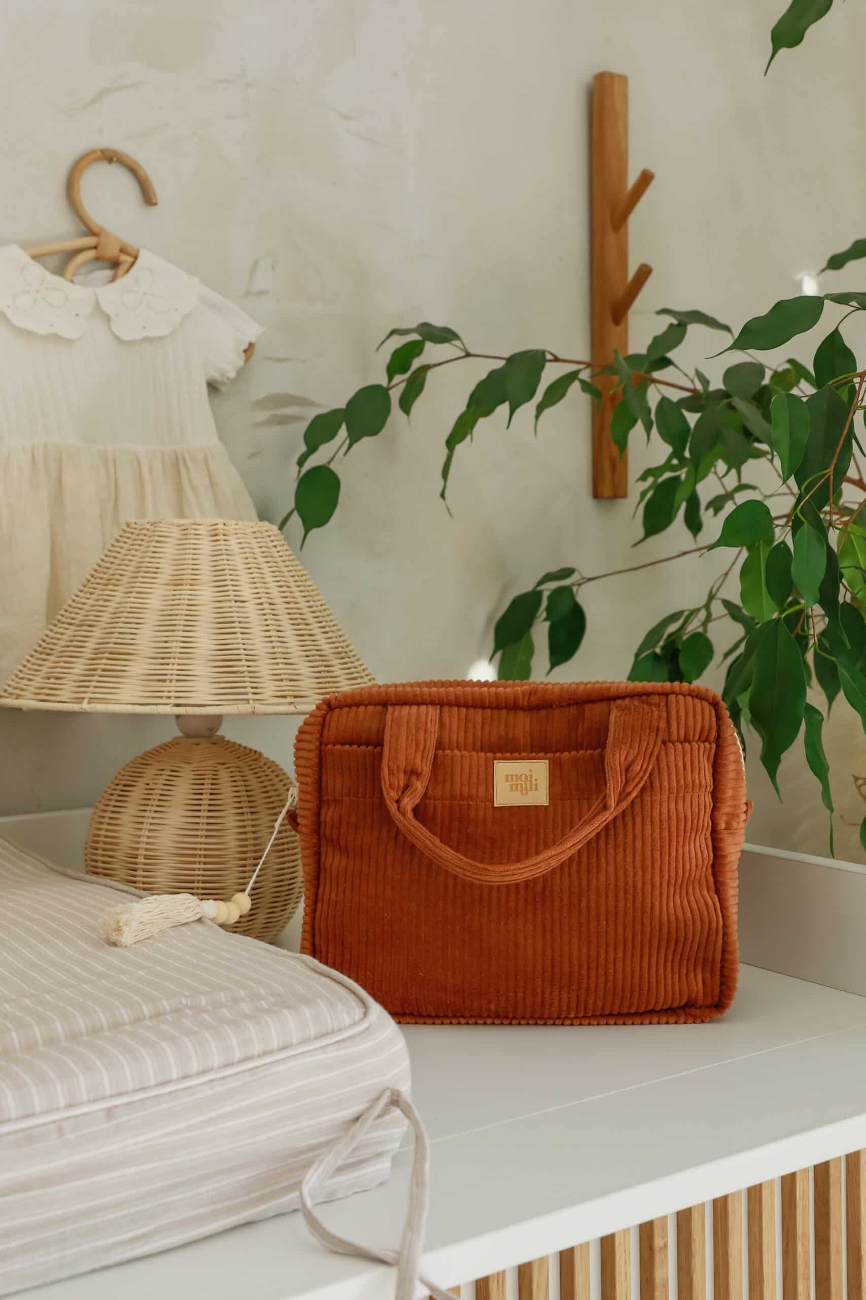 Brązowa torba na białym blacie obok wiklinowej lampy i białej sukienki na wieszaku w jasnym wnętrzu