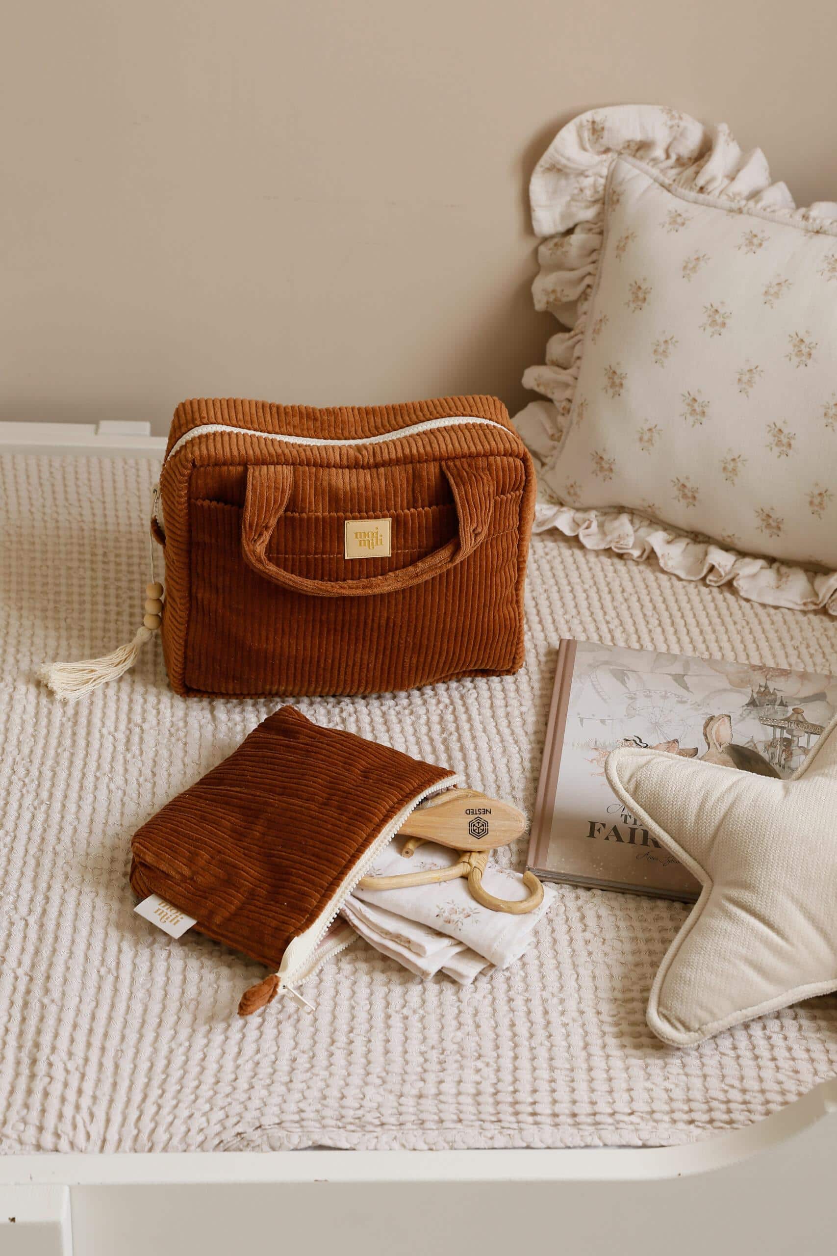 Brązowa wyszywana kosmetyczka i etui na jasnym plecionym kocu obok poduszki w kształcie gwiazdy i książki