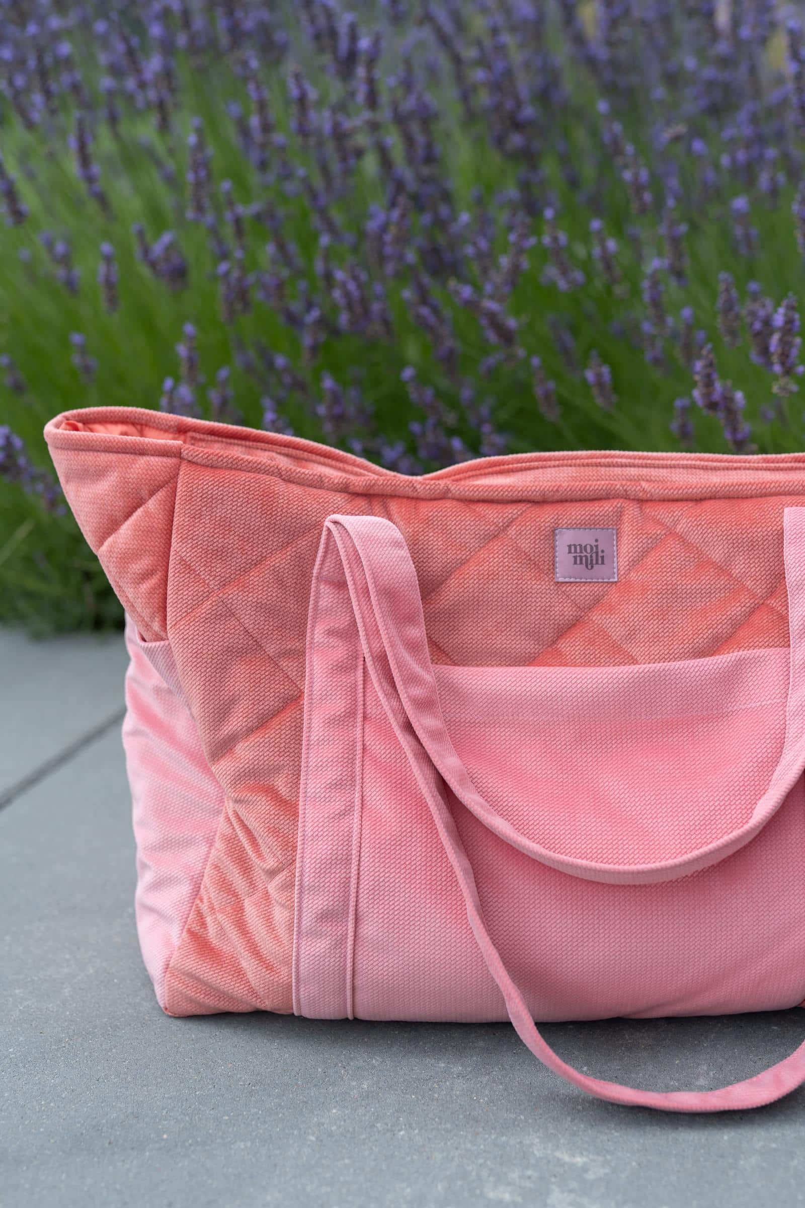 Różowa torba z przeszyciami na szarym chodniku na tle fioletowych kwiatów lawendy.