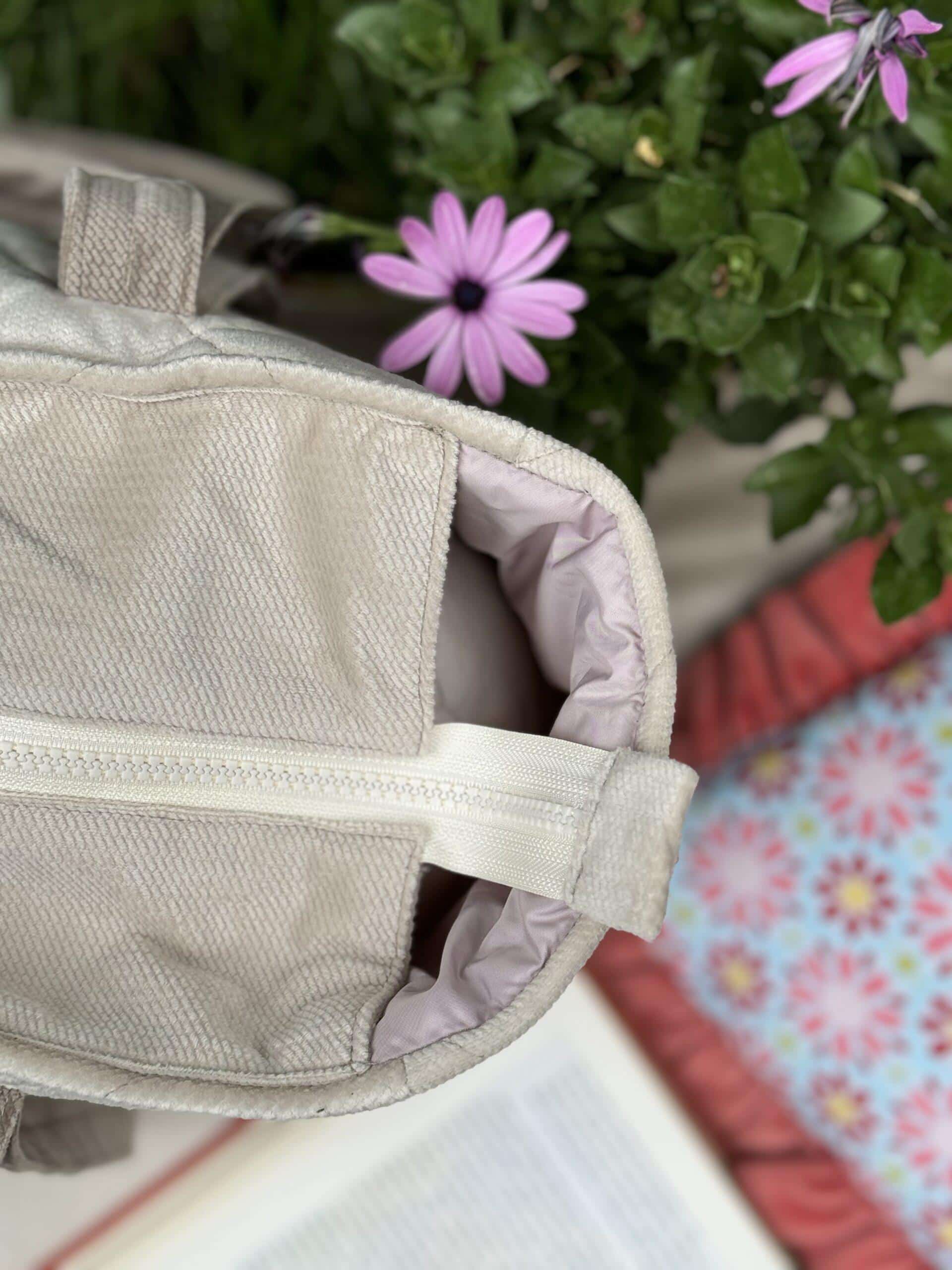 Zbliżenie na jasnoszarą kieszeń z zamkiem błyskawicznym na tle zielonych liści i fioletowych kwiatów