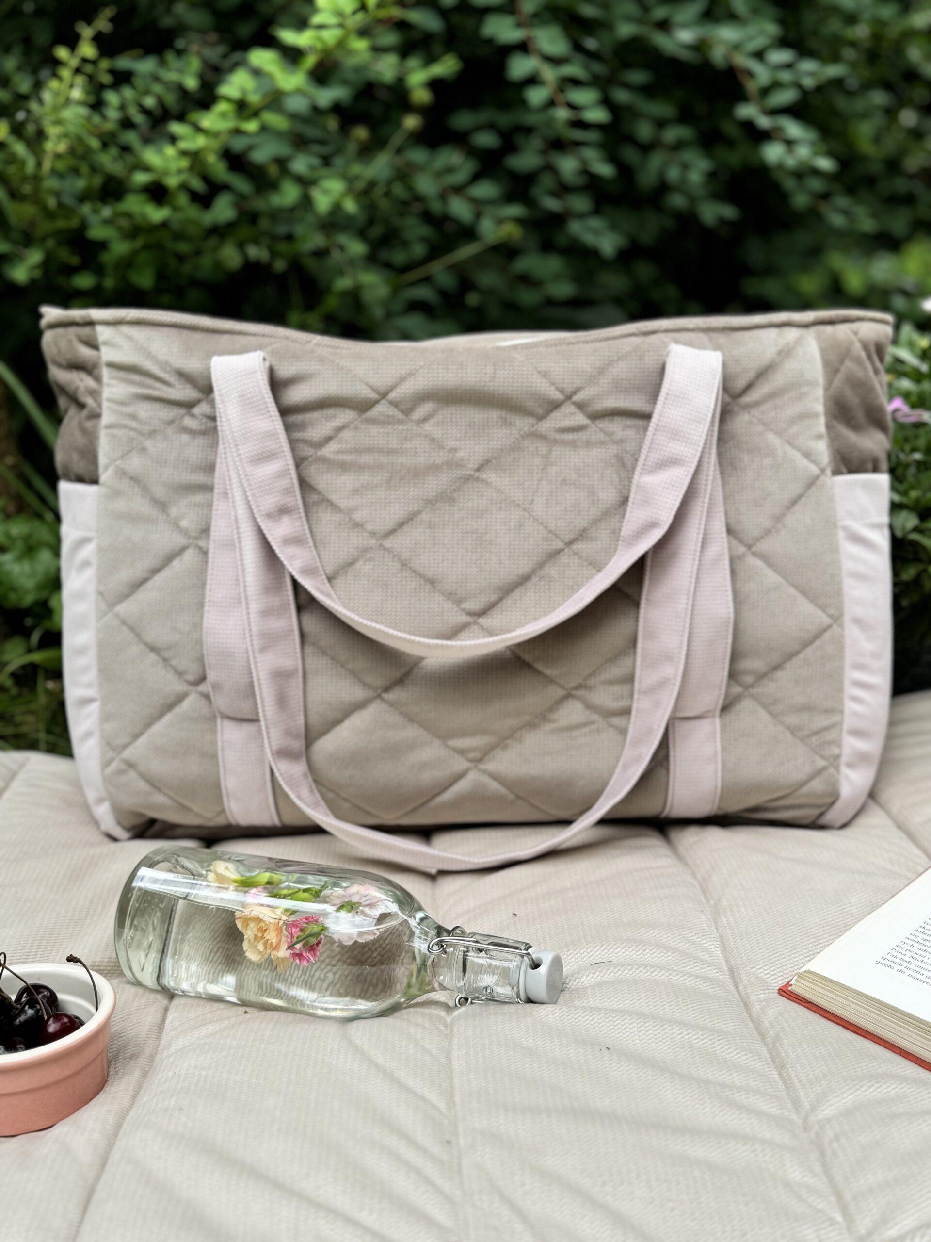 Pikowana torba na ramię w beżowych odcieniach na kołderce w ogrodzie, obok butelka z kwiatami i książka.