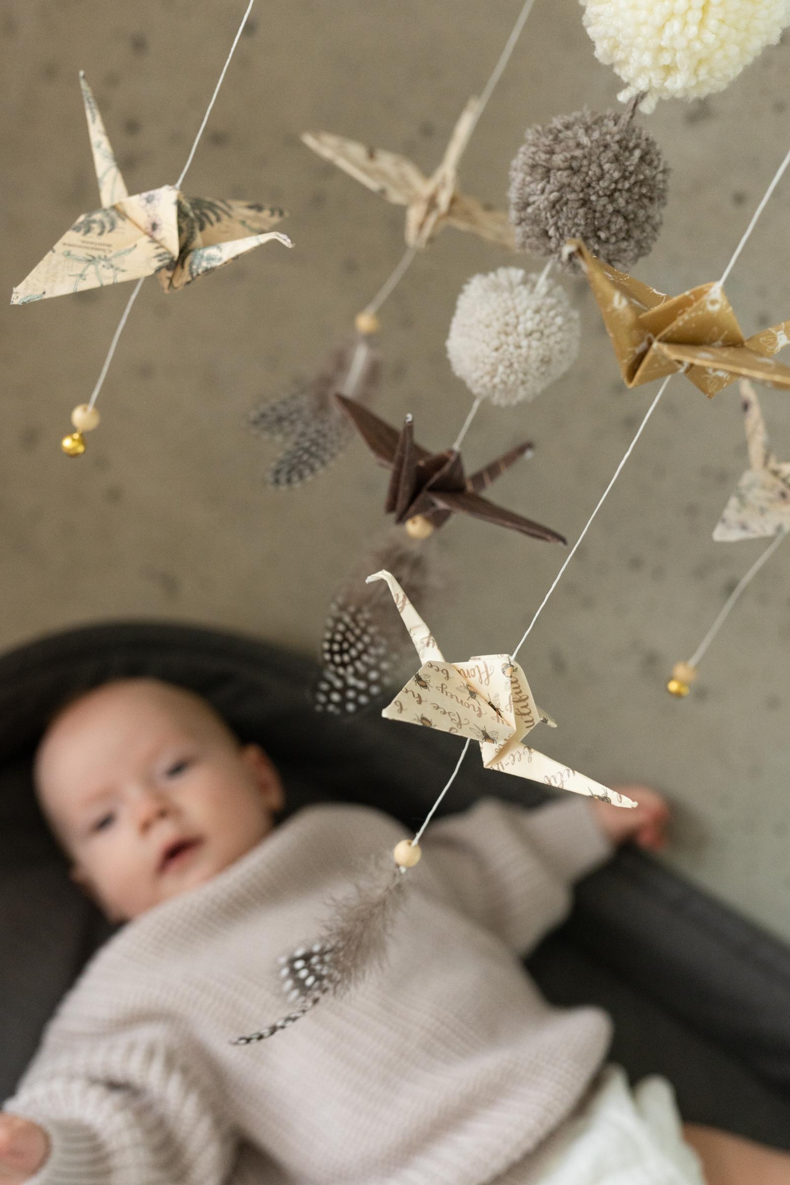 Mobil z papierowymi żurawiami i pomponami nad leżącym niemowlęciem w jasnym swetrze