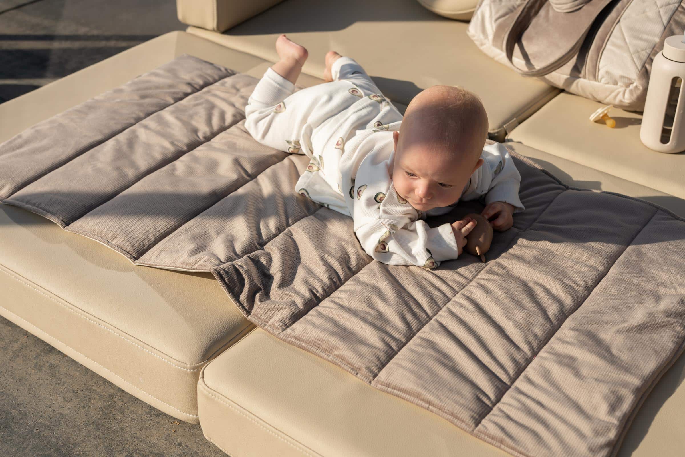 Niemowlę leży na szarobeżowej macie na jasnobeżowej kanapie, ubrany w białe ubranko z awokado