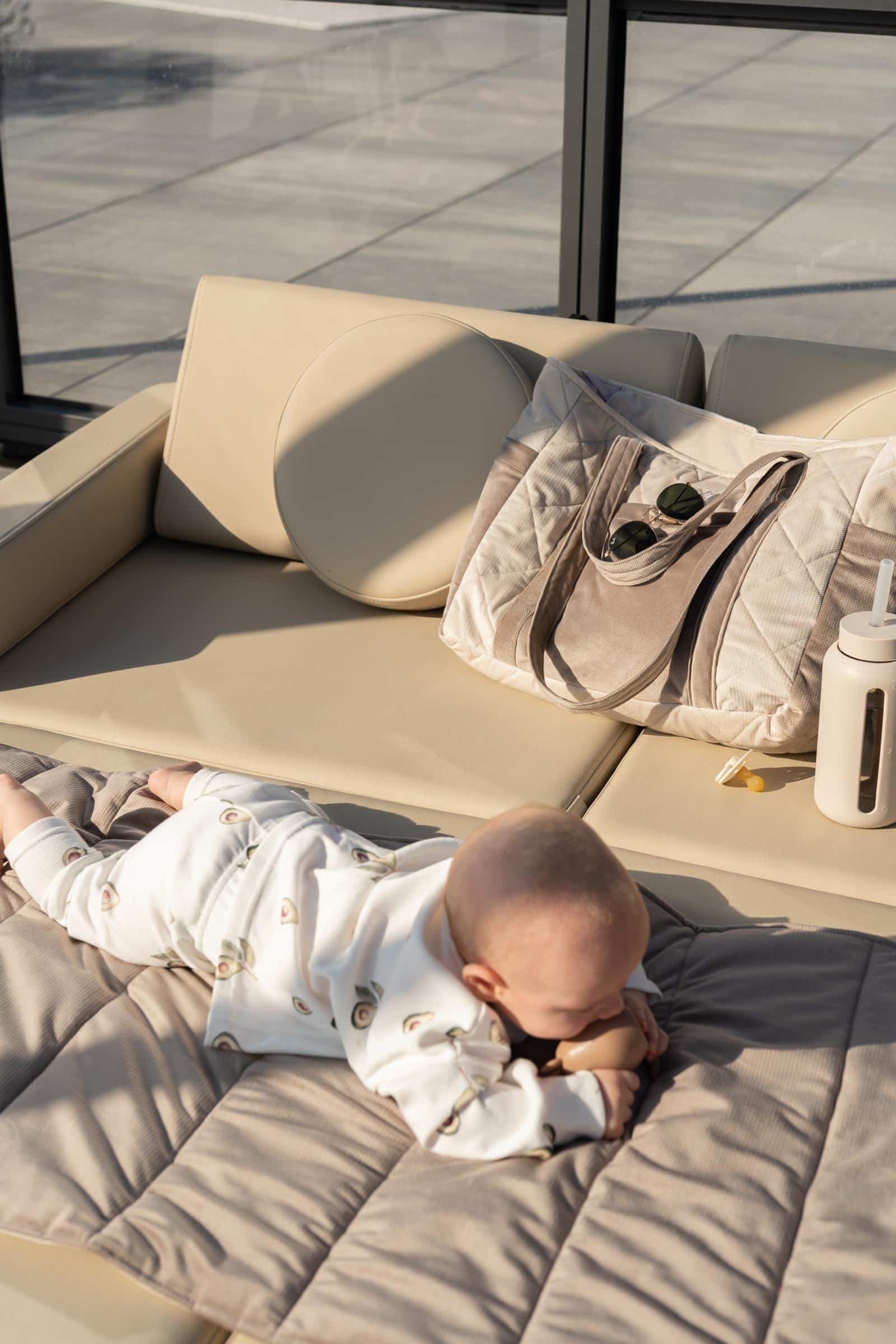 Niemowlę leżące na miękkim kocyku na beżowej sofie obok torby, butelki i smoczka w słońcu.