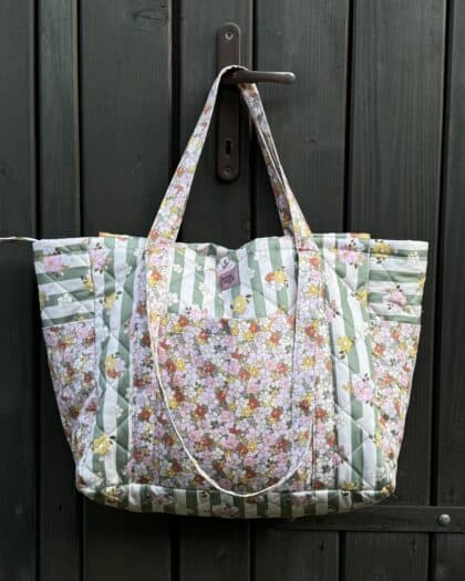 Pikowana torba z długimi uszami wisząca na metalowej klamce drzwi drewnianych w ciemnym kolorze, ozdobiona wzorem w zielone paski i kwiaty w pastelowych kolorach.