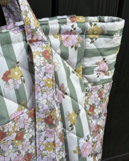 Zbliżenie na pikowaną torbę z zielono-białymi paskami i wzorem kolorowych kwiatów w odcieniach różu, żółci i pomarańczu, na tle ciemnej drewnianej powierzchni.