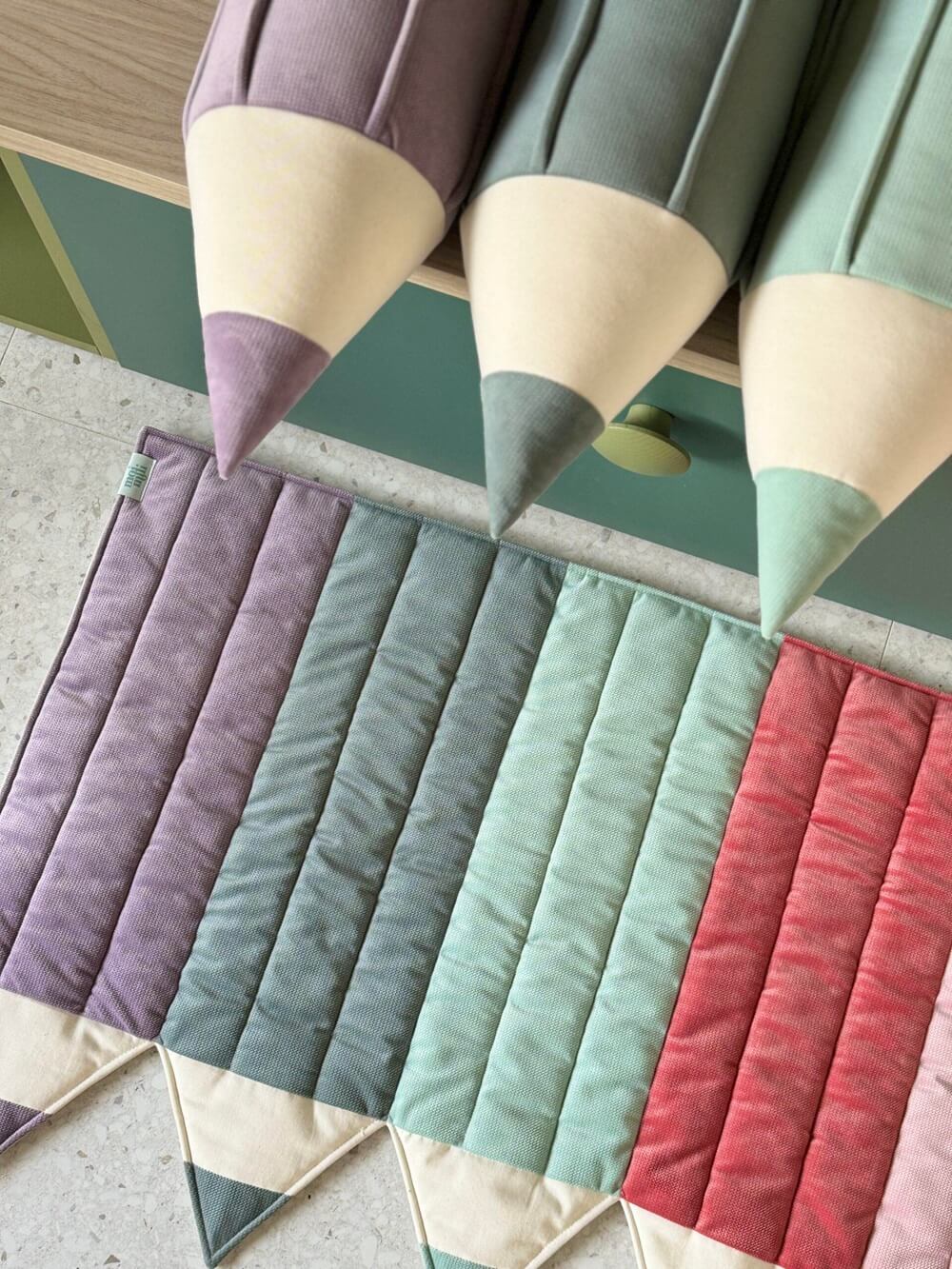 Trzy duże poduszki ołówki i kolorowa pikowana mata z pasami w odcieniach fioletu, zieleni i czerwieni.