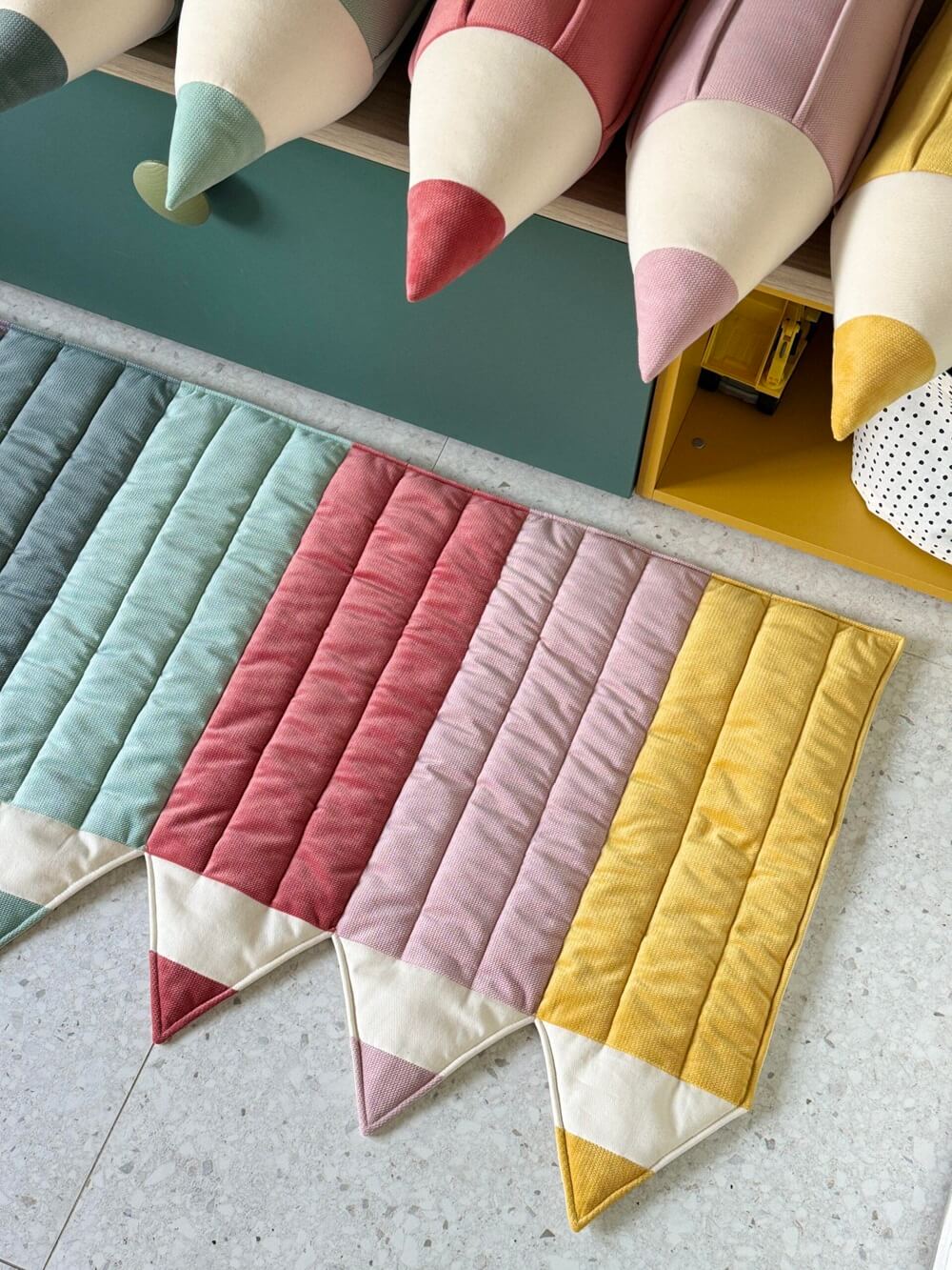 Kolorowe poduszki i dywanik w kształcie dużych kredek ustawione na podłodze i półce w pokoju dziecięcym