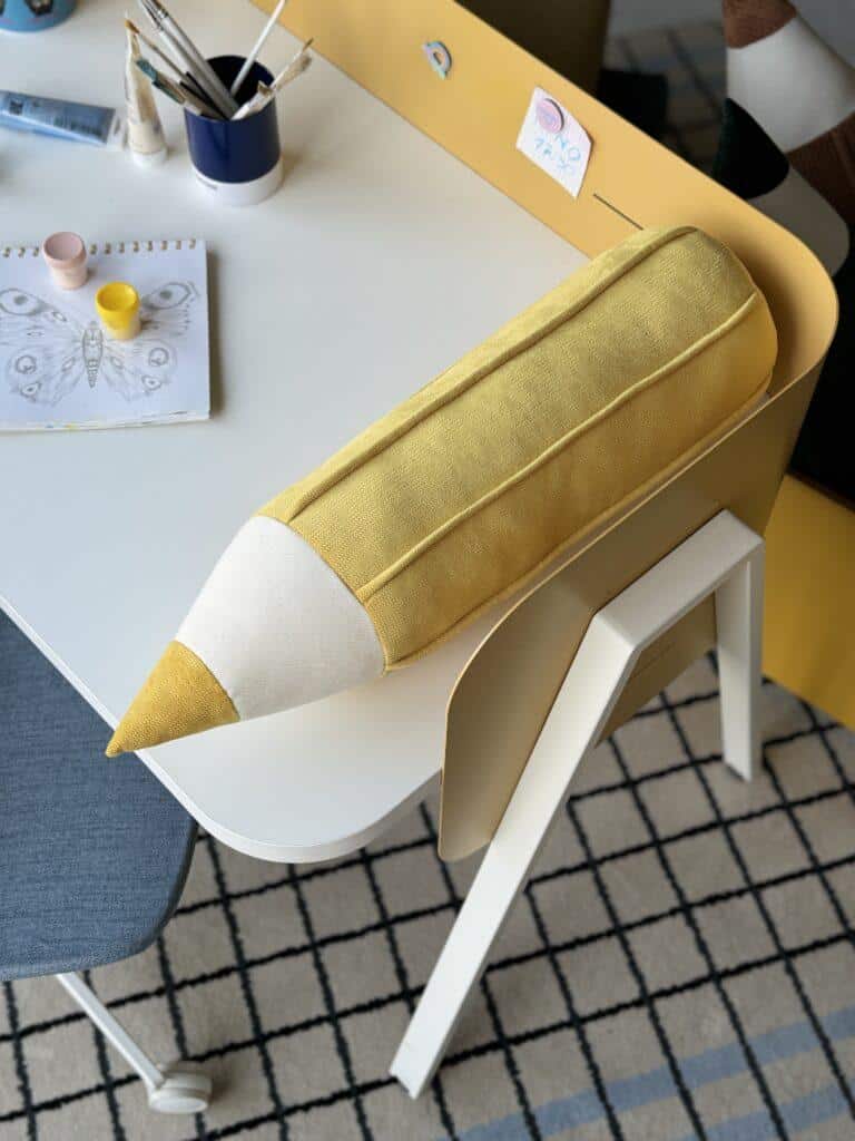 Żółta poduszka w kształcie ołówka na białym biurku z przyborami do malowania i szkicem motyla
