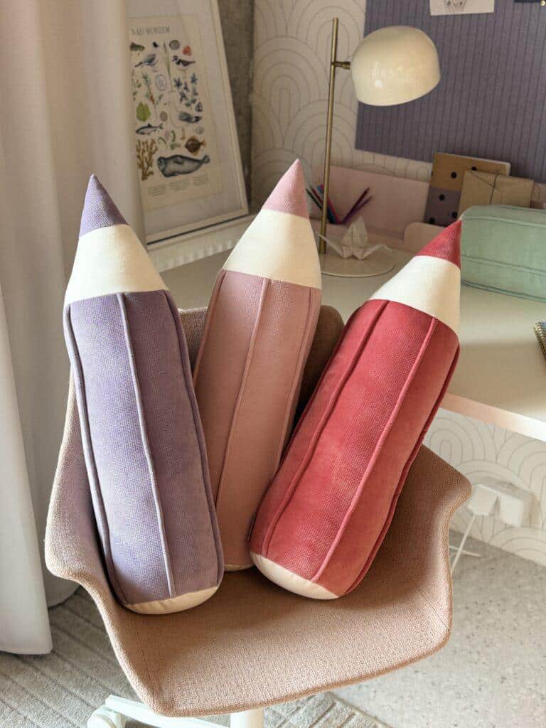 Trzy dekoracyjne poduszki na krześle w kształcie kolorowych kredek: fioletowa, różowa i czerwona.