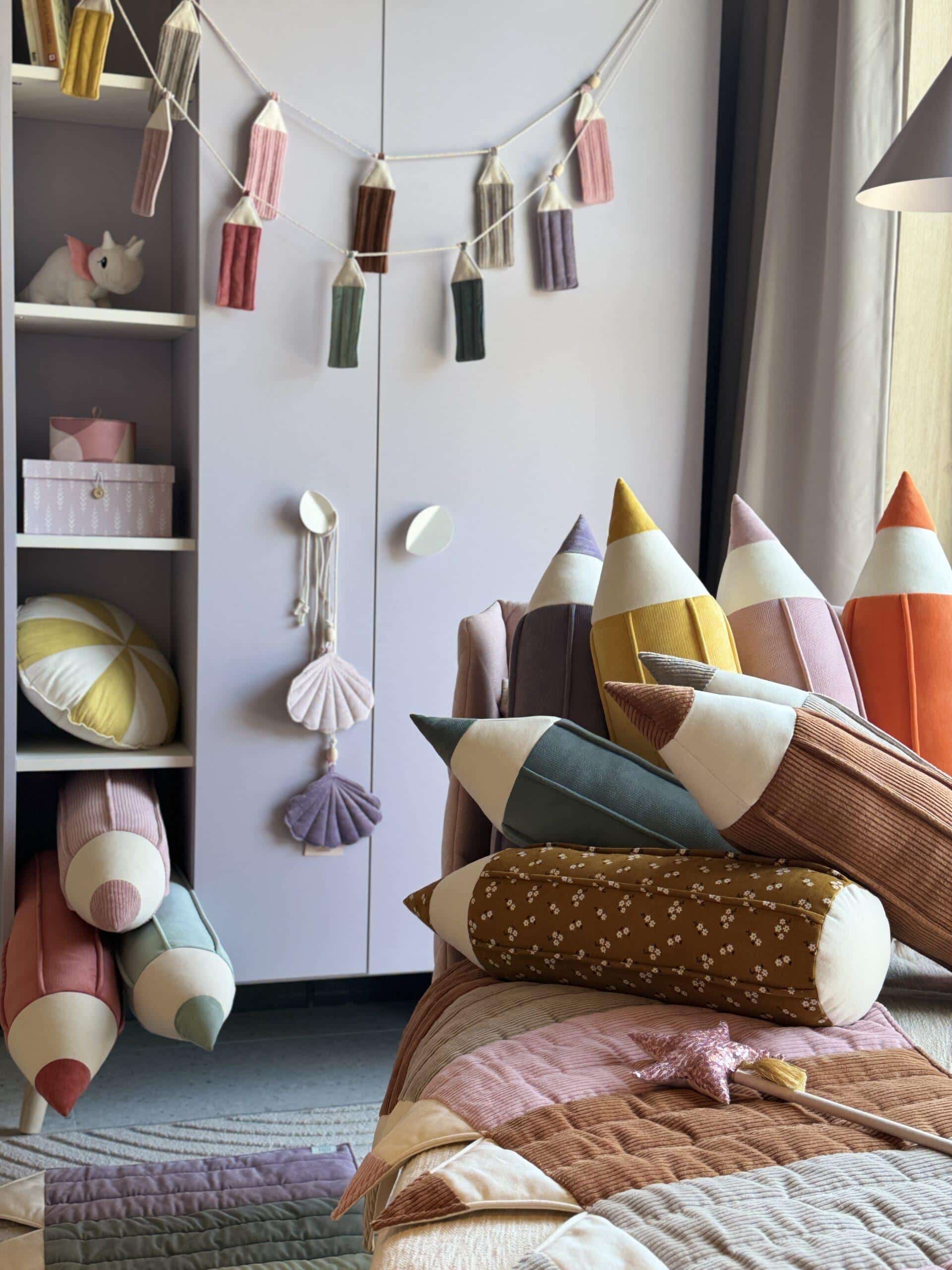 Pokój dziecięcy z kolorowymi poduszkami w kształcie kredek i dekoracjami na ścianie oraz półce