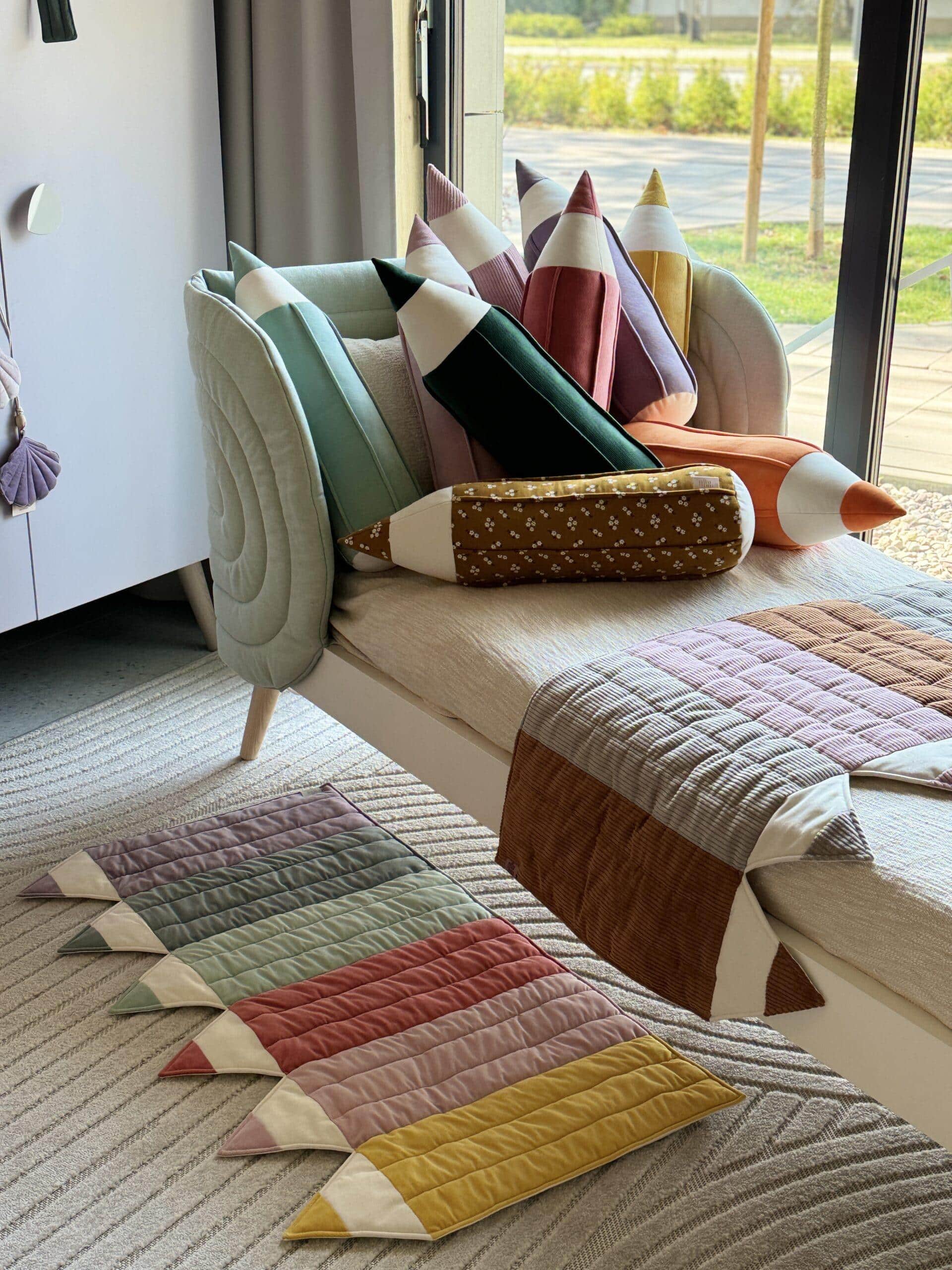 Łóżko z kolorowymi poduszkami w kształcie kredek i dopasowanym dywanikiem oraz narzutą w jasnym pokoju