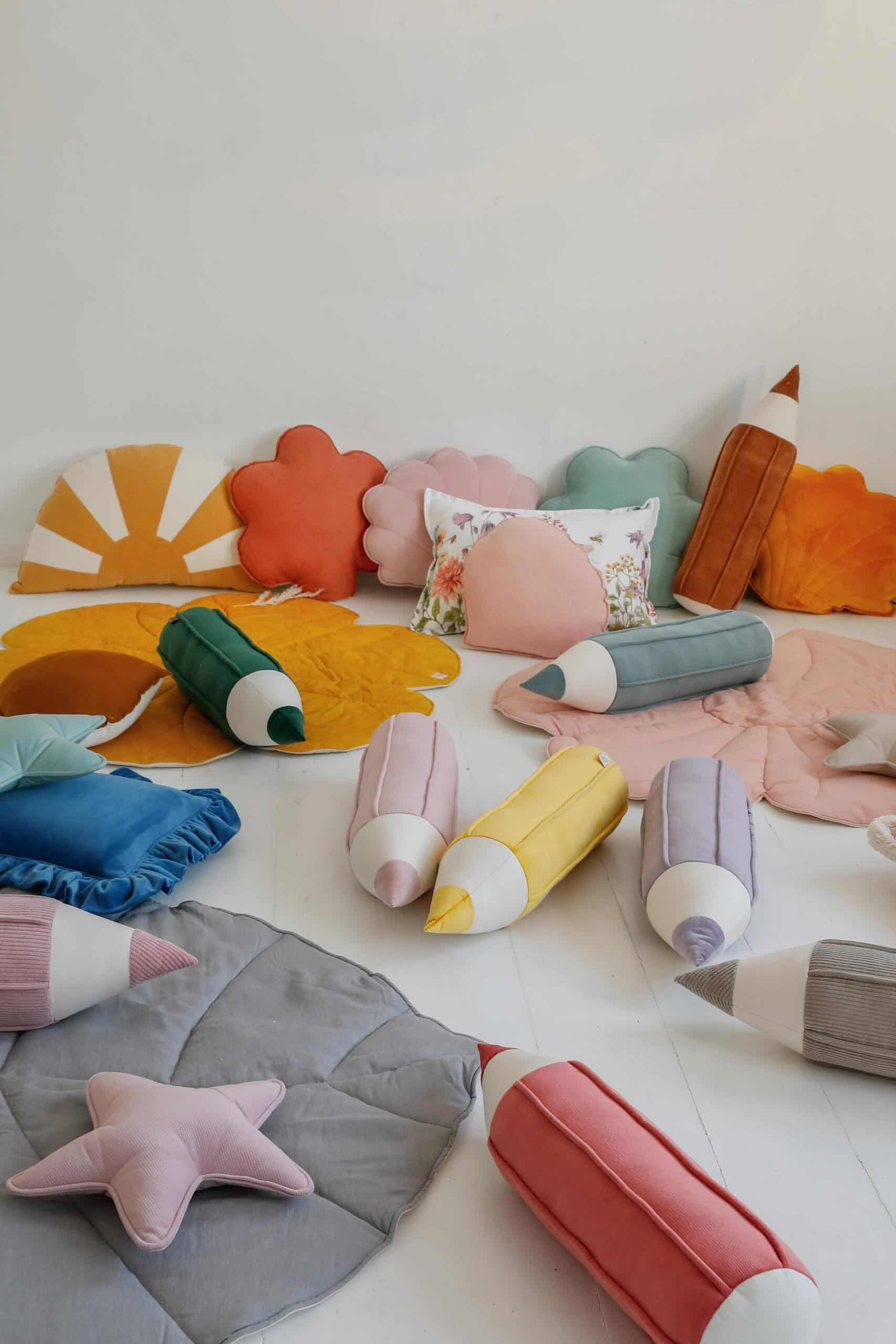 Kolorowe pluszowe poduszki i kształty w formie kredek oraz kwiatów rozmieszczone na jasnej podłodze w pokoju.