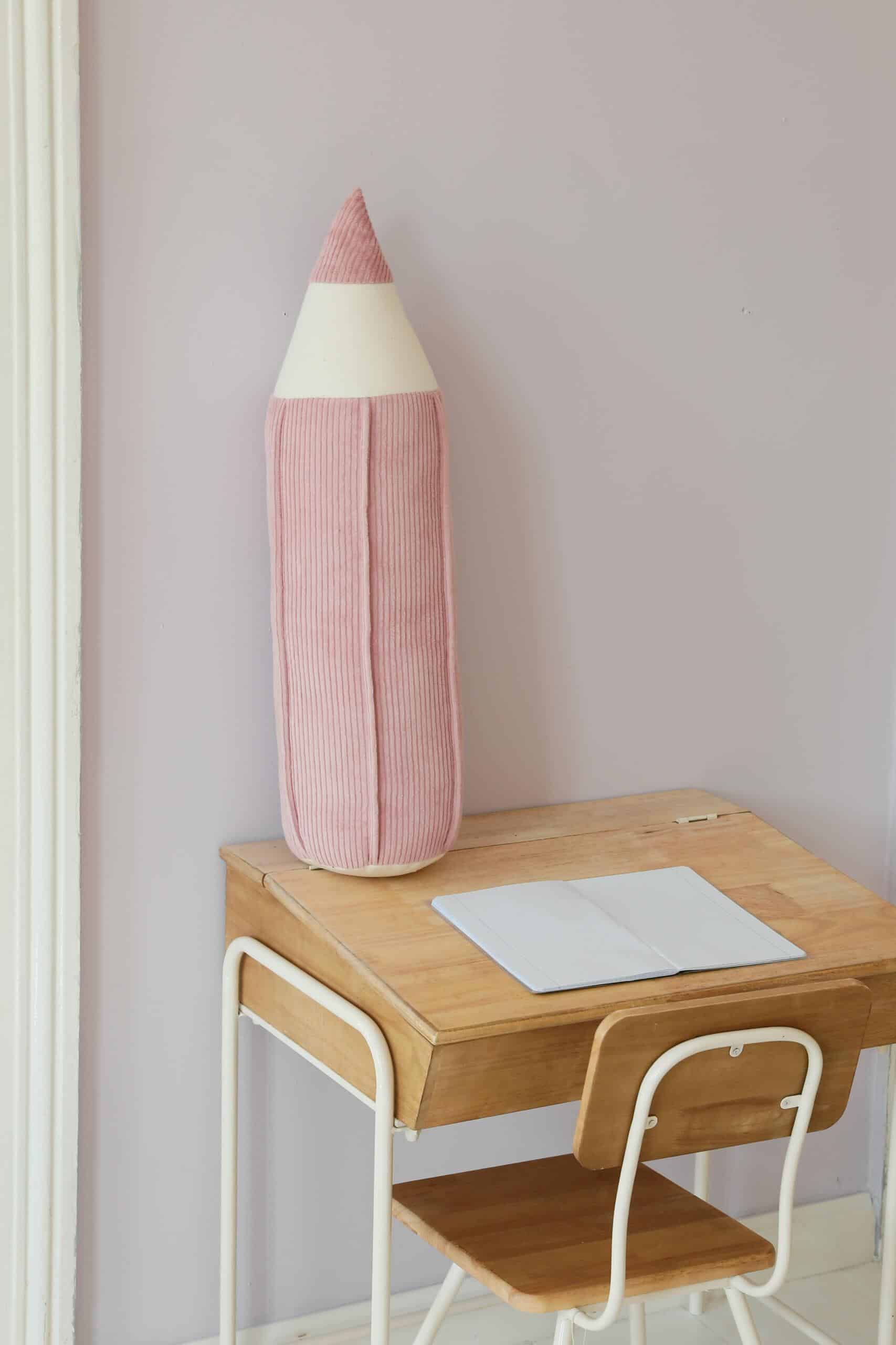 Małe drewniane biurko z krzesłem i różową poduszką w kształcie ołówka na jasnofioletowej ścianie