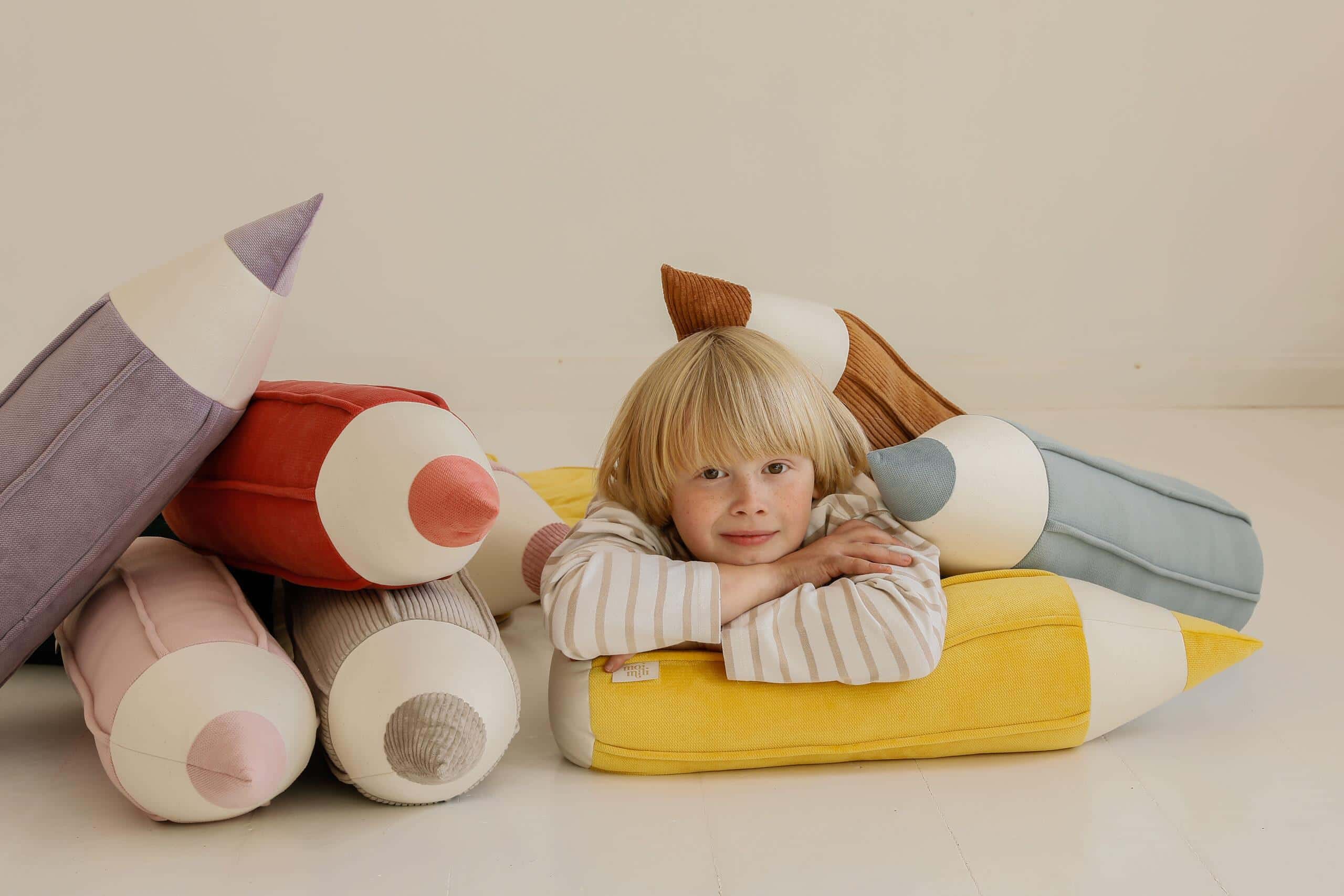 Dziecko leżące na podłożu z poduszkami w kształcie dużych kredek w pastelowych kolorach