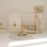 Drewniane łóżko dziecięce w kształcie domu z beżowym namiotem i poduszkami, jasne, minimalistyczne wnętrze