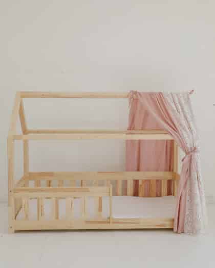 Drewniane łóżko dziecięce w kształcie domu z różowymi i białymi zasłonami na jasnym tle.