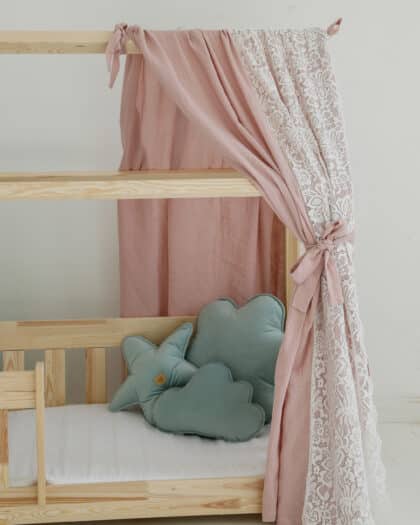 Drewniane łóżeczko dziecięce z zasłonami w różowym i koronkowym kolorze oraz poduszkami w kształcie chmury i gwiazdy.