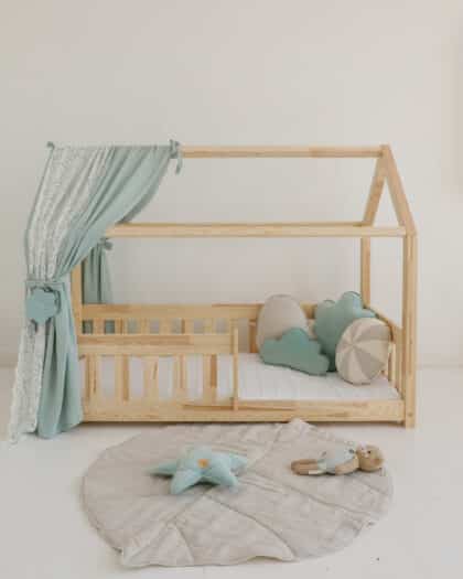 Drewniane łóżko dziecięce w kształcie domku z niebiesko-beżowymi poduszkami i zabawkami na jasnej podłodze