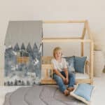 Chłopiec siedzi obok drewnianego łóżka-domku z niebieskimi poduszkami i szarą matą na podłodze