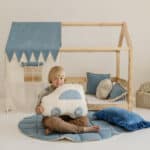 Chłopiec siedzi na niebieskim dywanie, trzyma poduszkę w kształcie samochodu przed drewnianym łóżkiem z baldachimem.