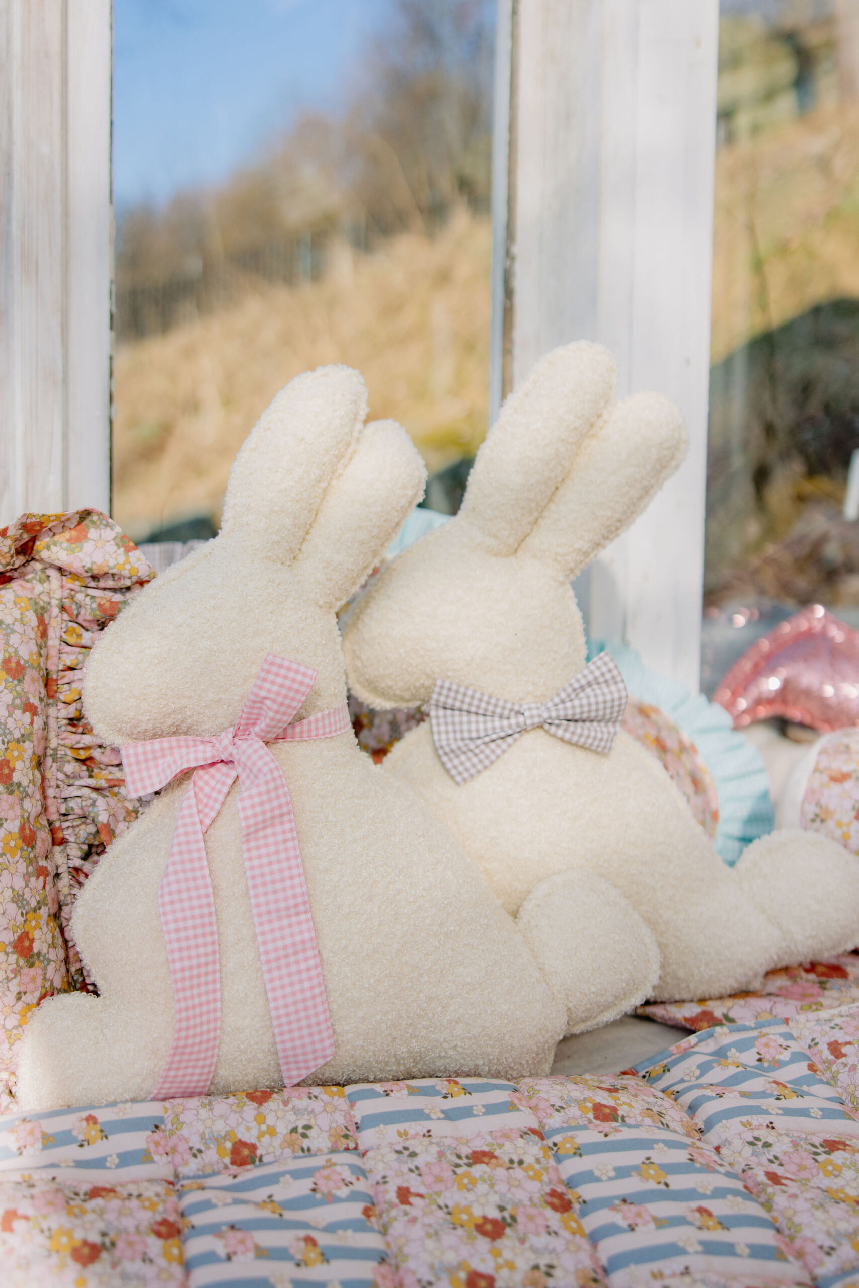 Dwa miękkie białe pluszowe zające z kokardkami, siedzące na patchworkowej narzucie przy oknie.